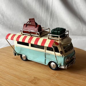 A vintage toy camper van, teal and cream with roof luggage and striped chairs, sits on a wooden surface.