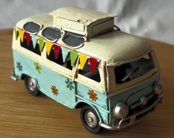 May include: A miniature, vintage-style camper van in shades of light blue and cream. The van features flower decals, a decorative banner, and a pop-up roof. The wheels are black, and the van appears to be made of metal.