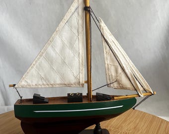 May include: A wooden model sailboat with a green hull and brown base. The sails are white, and the mast is a light brown colour. The boat is a decorative item, suitable for nautical-themed decor.