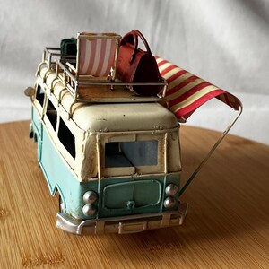 A vintage toy camper van, teal and cream with roof luggage and striped chairs, sits on a wooden surface.