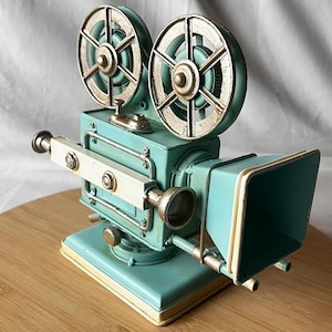 May include: A vintage-style teal and cream-colored metal movie camera. The camera features two large film reels, a lens, and a rectangular viewfinder. The decorative piece is set on a wooden surface.