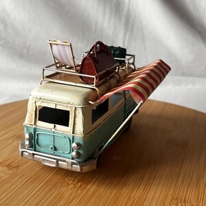 A vintage toy camper van, teal and cream with roof luggage and striped chairs, sits on a wooden surface.
