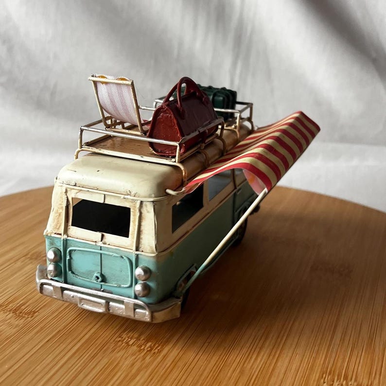 A vintage toy camper van, teal and cream with roof luggage and striped chairs, sits on a wooden surface.