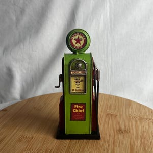 Puede incluir: Una bomba de gasolina decorativa de metal verde y rojo de estilo vintage. Tiene la palabra "GASOLINE" en la parte superior y frontal, con una estrella roja y las palabras "Fire Chief".