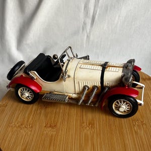 Può includere: Un'auto giocattolo vintage color crema con parafanghi rossi e pneumatici neri. L'auto presenta interni neri, una ruota di scorta e dettagli in metallo color argento. L'auto è esposta su una superficie di legno.