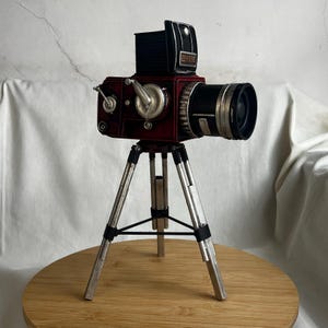 May include: A vintage-style red metal camera on a silver tripod. The camera has a black lens and a black and silver body. The camera is on a wooden surface.