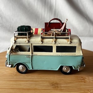 A vintage toy camper van, teal and cream with roof luggage and striped chairs, sits on a wooden surface.