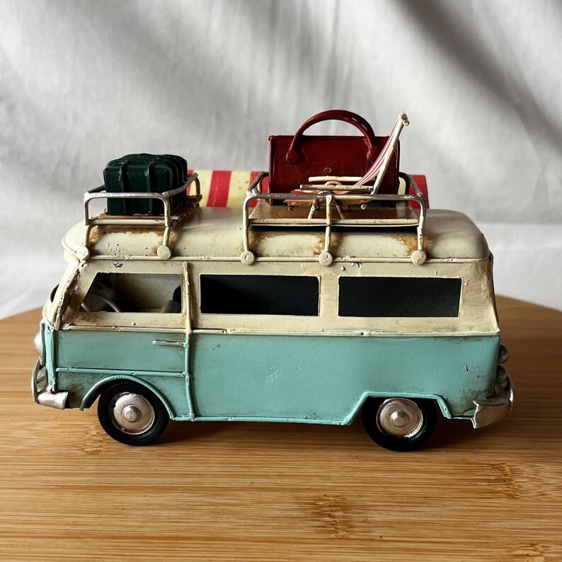 A vintage toy camper van, teal and cream with roof luggage and striped chairs, sits on a wooden surface.