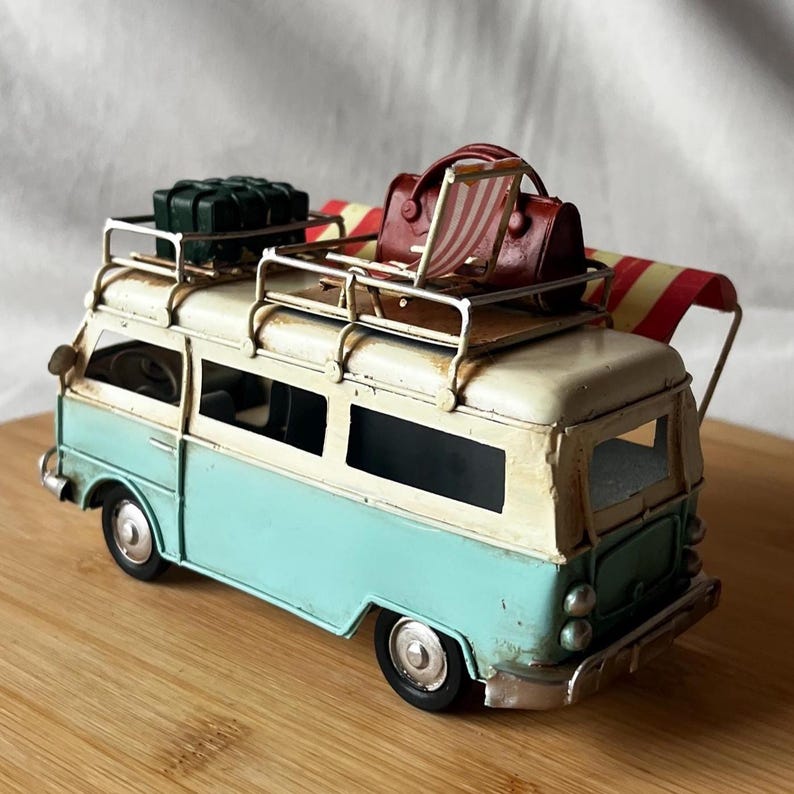 A vintage toy camper van, teal and cream with roof luggage and striped chairs, sits on a wooden surface.