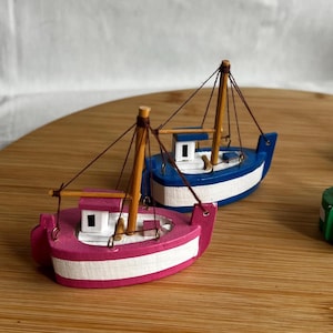 Puede incluir: Dos pequeños barcos de juguete de madera. Uno está pintado de rosa y blanco, el otro de azul y blanco. Ambos tienen mástiles y aparejos, y están sobre una superficie de madera.