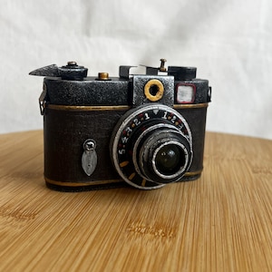 May include: A black and silver vintage-style camera with a gold trim and a lens with a black and silver ring. The camera has a black leather body and a silver metal lens.