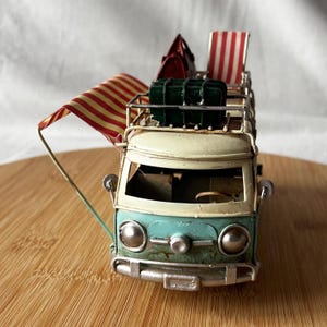 A vintage toy camper van, teal and cream with roof luggage and striped chairs, sits on a wooden surface.