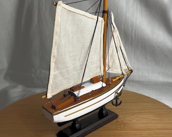 May include: A model sailing boat featuring a white hull with a thin gold stripe, a wooden deck, and two off-white fabric sails attached to a central mast. The vessel is secured to a dark rectangular wooden base and includes a small black anchor.