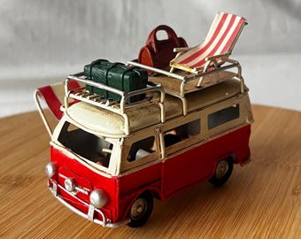 May include: A miniature red and cream-colored vintage van with luggage, a beach chair, and a surfboard on top. The van has a weathered appearance, suggesting a well-loved travel theme. The beach chair has red and white stripes.