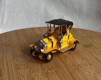 May include: A vintage yellow toy car with a black roof and silver headlights. The miniature vehicle has four black wheels and is set against a light background. The car's design is reminiscent of classic automobiles.