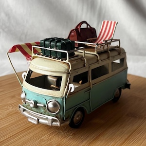 May include: A miniature teal and cream-coloured van with a luggage rack. The rack carries a green suitcase, a maroon bag, and a red and white striped deckchair. A red and yellow striped awning extends from the side.
