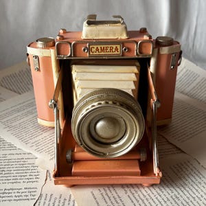 May include: A vintage-style decorative camera in a peach color with a gold-colored lens. The word "CAMERA" is displayed on the top. The camera is made of metal and has a classic, retro design, sitting on a background of printed paper.
