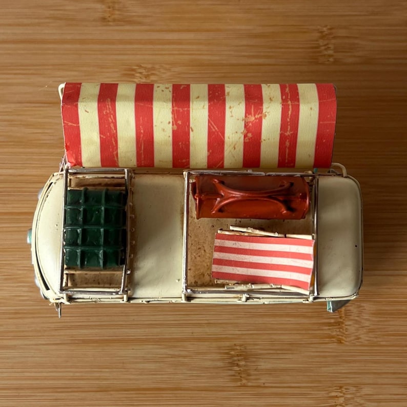 A vintage toy camper van, teal and cream with roof luggage and striped chairs, sits on a wooden surface.