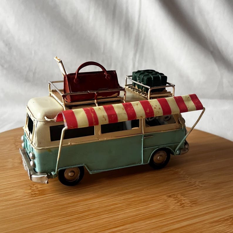 A vintage toy camper van, teal and cream with roof luggage and striped chairs, sits on a wooden surface.