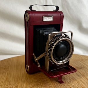 Puede incluir: Una cámara de metal roja de estilo vintage con una lente negra y un engranaje plateado. La cámara está abierta y la lente está orientada hacia el espectador.