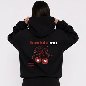 Puede incluir: Una sudadera negra con capucha y un gráfico rojo en la parte posterior. El diseño incluye el texto lambda mu, la frase with love y un dibujo de dos cerezas atadas con un lazo. Las letras A y M están dentro de las cerezas, y el texto LIMITED EDITION aparece debajo.
