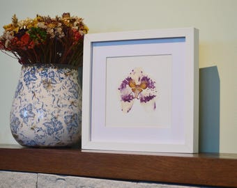 May include: White framed art print featuring a purple and yellow orchid flower. A blue and white patterned vase with dried flowers sits to the left. The artwork is displayed on a dark brown shelf.