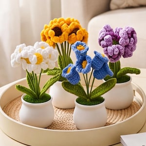 May include: Four small potted plants featuring crocheted flowers in white, yellow, blue, and purple. Each plant has green crocheted leaves and sits in a small white ceramic pot. The four pots are arranged together on a round, light-colored tray.