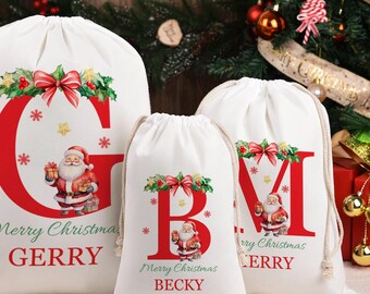 May include: Three white drawstring Christmas gift bags with red letters, Santa Claus illustrations, and the text "Merry Christmas". The bags are decorated with bows and holly. The largest bag has the letter "G" and the name "Gerry".