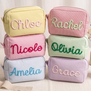 May include: Six rectangular zippered pouches made of seersucker fabric in yellow, pink, light pink, green, light blue, and lavender. Each pouch features a name embroidered in a cursive font: Chloe, Rachel, Nicole, Olivia, Amelia, and Grace.
