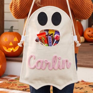 Puede incluir: Una bolsa de lona color crema con forma de fantasma para dulces, con dos ojos de fieltro negro y una abertura central llena de golosinas. El nombre Carlin est&aacute; bordado en hilo rosa en la parte frontal, con asas de cuerda blanca.