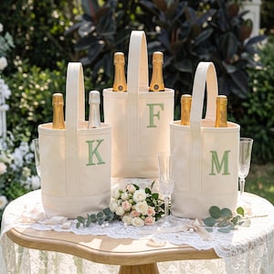 May include: Three off-white canvas wine tote bags with single vertical handles. Each bag features a large, embroidered sage green letter on the front: K, F, and M. Each bag holds two bottles of champagne with gold foil labels and necks visible.