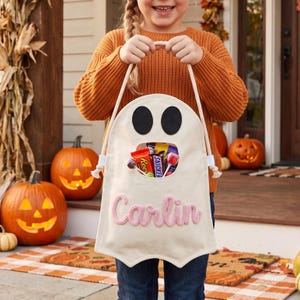 Puede incluir: Una bolsa de lona color crema con forma de fantasma para dulces, con dos ojos de fieltro negro y una abertura central llena de golosinas. El nombre Carlin est&aacute; bordado en hilo rosa en la parte frontal, con asas de cuerda blanca.