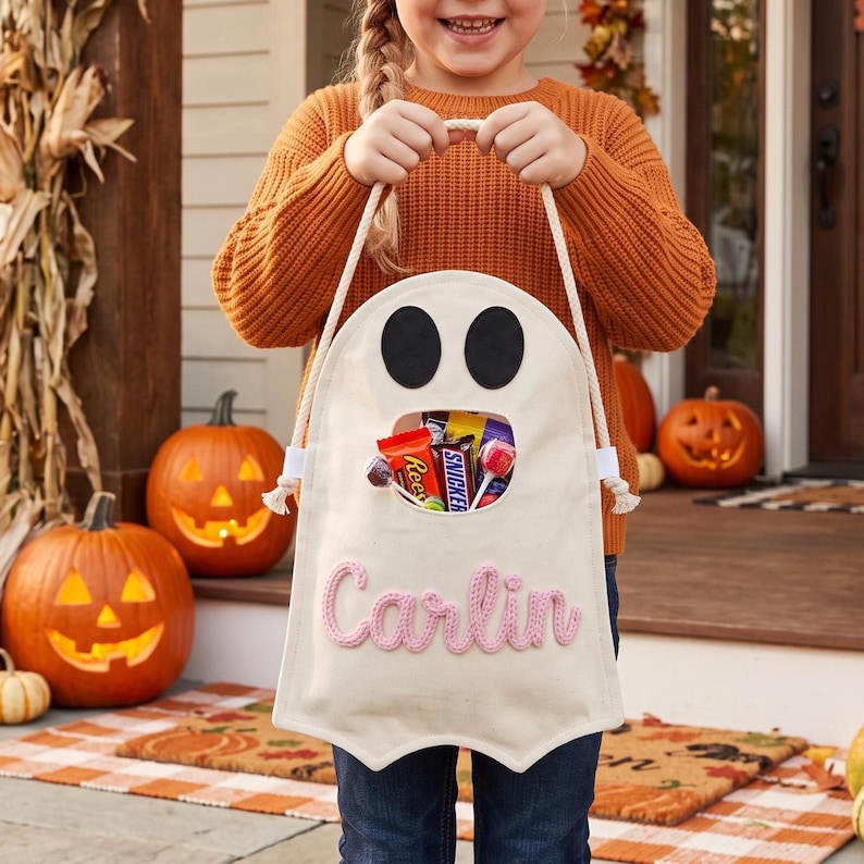 Puede incluir: Una bolsa de lona color crema con forma de fantasma para dulces, con dos ojos de fieltro negro y una abertura central llena de golosinas. El nombre Carlin est&aacute; bordado en hilo rosa en la parte frontal, con asas de cuerda blanca.