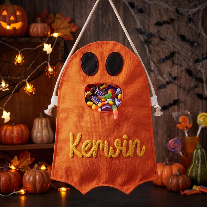 Puede incluir: Una bolsa de tela naranja con forma de fantasma que presenta dos ojos ovalados negros, una boca abierta central llena de dulces variados y el nombre Kerwin bordado en hilo amarillo. La bolsa cuelga de un asa de cuerda blanca gruesa.