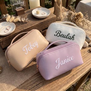 May include: Three rectangular cosmetic bags with top handles and zipper closures. One bag is beige with the name Tobey in white, one is white with the name Beulah in black, and one is lavender with the name Janice in white script lettering.