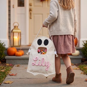 Puede incluir: Una bolsa de tela color crema con forma de fantasma, con dos ojos de fieltro negro, una abertura circular llena de dulces variados y el nombre Lily bordado en hilo rosa en la parte frontal, sujeta por un asa de cuerda blanca gruesa.
