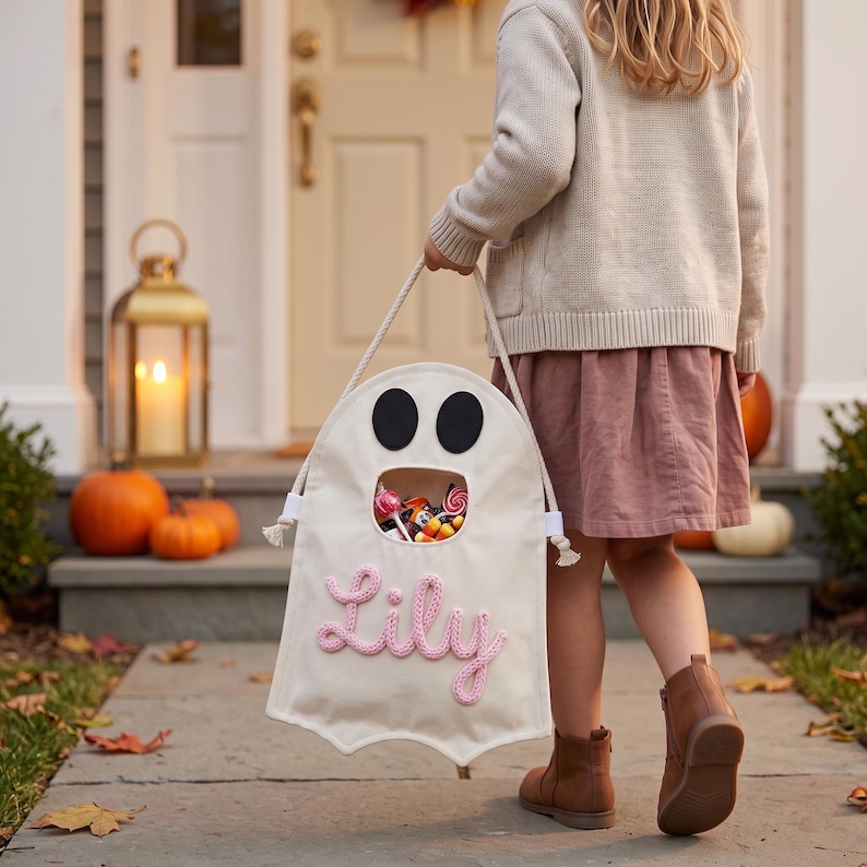 Puede incluir: Una bolsa de tela color crema con forma de fantasma, con dos ojos de fieltro negro, una abertura circular llena de dulces variados y el nombre Lily bordado en hilo rosa en la parte frontal, sujeta por un asa de cuerda blanca gruesa.