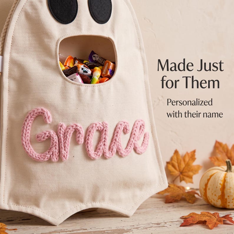 Puede incluir: Una bolsa de tela color crema con forma de fantasma, dos ojos de fieltro negro y una abertura central llena de dulces. El nombre Grace est&aacute; bordado con hilo rosa en la parte frontal. El texto dice: Made Just for Them. Personalized with their name.