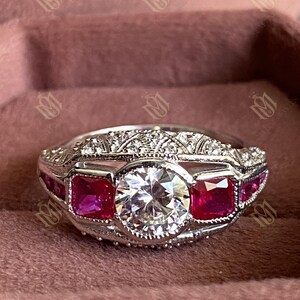 Pode incluir: Um anel de prata com uma grande pedra central redonda e transparente, ladeada por duas pedras vermelhas quadradas. O anel tem detalhes intrincados ao longo da banda e ao redor das pedras, sobre um fundo rosa. Uma joia.