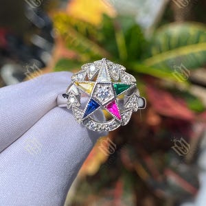 May include: A silver ring featuring a multi-colored star design. The star has a central diamond and is surrounded by smaller diamonds and colored enamel in yellow, blue, pink, green, and white. The ring is held against a blurred background of green and yellow foliage.