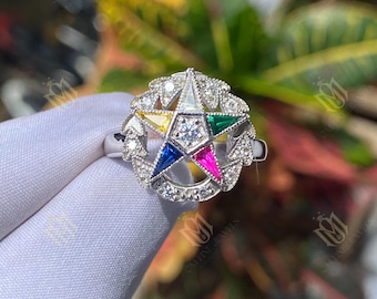 May include: A silver ring featuring a multi-colored star design. The star has a central diamond and is surrounded by smaller diamonds and colored enamel in yellow, blue, pink, green, and white. The ring is held against a blurred background of green and yellow foliage.