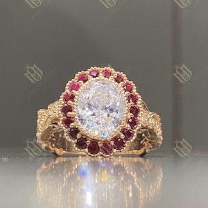 May include: A rose gold ring featuring a large, oval-cut, clear gemstone surrounded by a halo of smaller, round, red gemstones. The ring's band has an ornate, textured design.