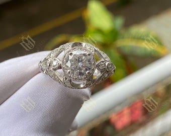 May include: A silver ring with a large, round, clear gemstone at the center. The ring has a detailed, ornate design with smaller accent stones and filigree work. The ring is displayed against a blurred green background.