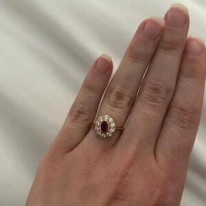 Pode incluir: Um anel de ouro com uma pedra vermelha oval central, rodeada por um halo de pequenas pedras claras. O anel é montado numa delicada banda de ouro, apresentando um design clássico e elegante.
