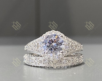 May include: A two-piece, silver-toned ring set. The engagement ring features a large, round, clear gemstone surrounded by smaller stones. The band is detailed with intricate patterns. The matching band has a similar design.