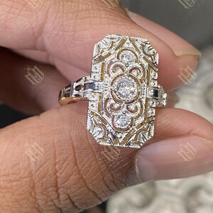 May include: A rectangular, Art Deco-style ring with a gold band and intricate filigree design. The ring features three round, clear gemstones set in a silver-colored metal. The filigree has a gold-colored background.