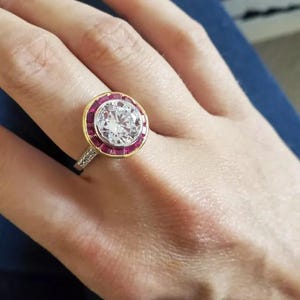 May include: A gold and silver ring with a large, clear center stone surrounded by a halo of small, red stones. The ring is set on a silver band with small accent stones. The ring is on a person's finger.