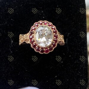 May include: A gold ring featuring a large, oval, clear gemstone encircled by small, red gems. The ring has an elaborate, floral design around the central stones and on the band. The ring is displayed against a black backdrop.