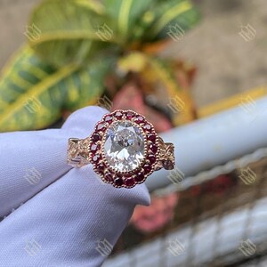 Puede incluir: Anillo de oro rosa con una gema ovalada y transparente, rodeada por un halo de pequeñas gemas rojas. La banda del anillo tiene detalles intrincados en los laterales. El anillo se muestra sobre una superficie blanca.