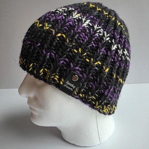 May include: A black knit beanie with a textured pattern, accented with purple, yellow, and white flecks. The beanie has a leather tag that reads "Spooky Artworks".
